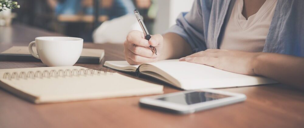 woman writing on a notebook beside teacup and tablet computer