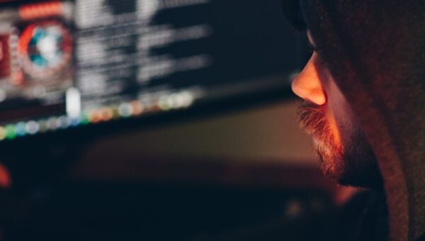 Side view of crop anonymous cyber spy in hoodie hacking computer system with information on screen at night