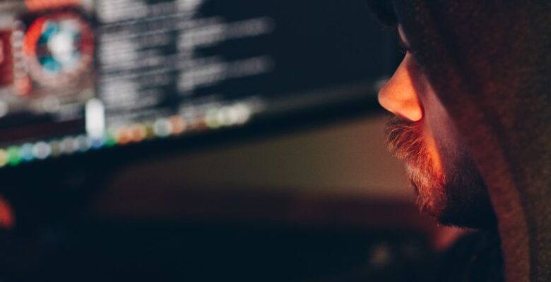 Side view of crop anonymous cyber spy in hoodie hacking computer system with information on screen at night